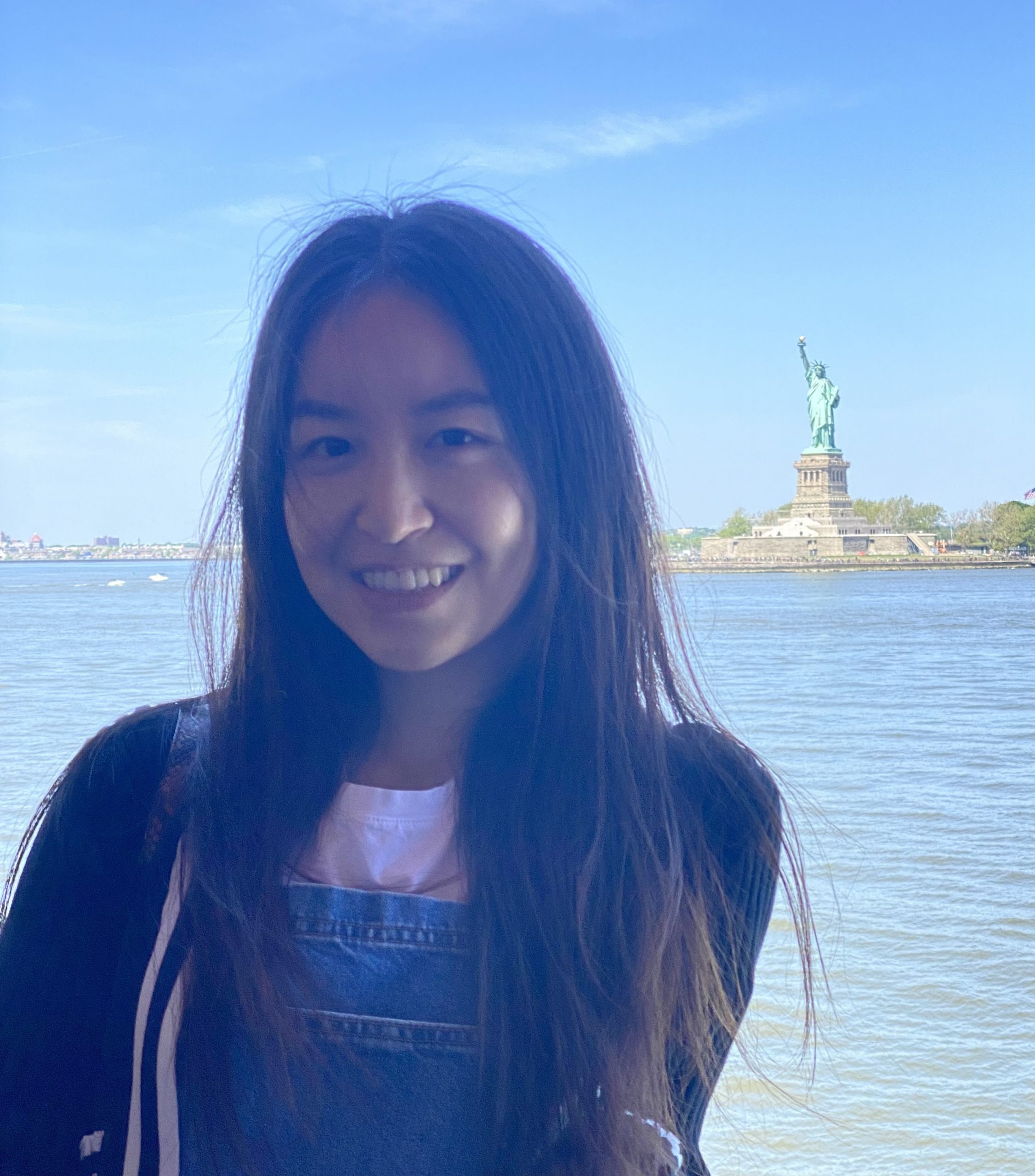 a young woman with dark hair stands posing with the statue of liberty in the background